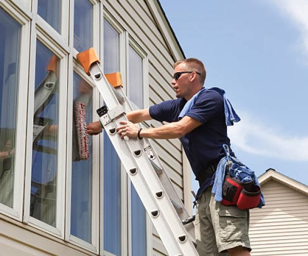 Young business owner washes windows. Window washing professional. Residential window is cleaned from a ladder with wetting wands, squeegee and cloths. Beautiful sunny day. Reflections show in the windows.